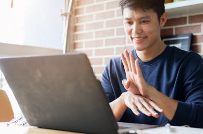 A smiling man uses sign language while video chatting with someone on his laptop.