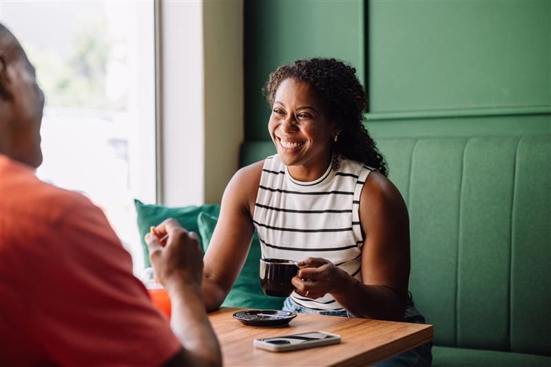 A woman smiling and holding a coffee cup while talking with someone across the table in a café. A smartphone and saucer are on the table in front of her.
