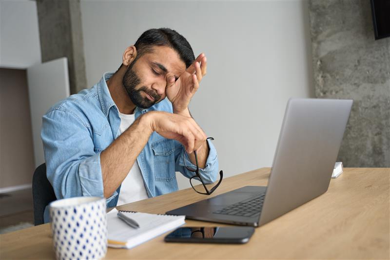 A man with dark hair and a beard, wearing a blue denim shirt, rubs his eye with one hand while holding a pair of glasses in the other, looking tired in front of an open laptop. 