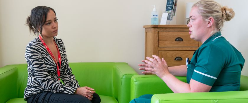 An occupational therapist wearing a zebra-print blouse and a veterinary nurse in a green uniform having a conversation in a healthcare setting while sitting on bright green chairs.