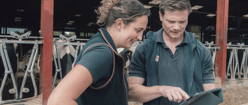 Two veterinary professionals, a woman and a man, stand in front of a barn looking at a tablet together. The woman is smiling, and cows are visible in the background.