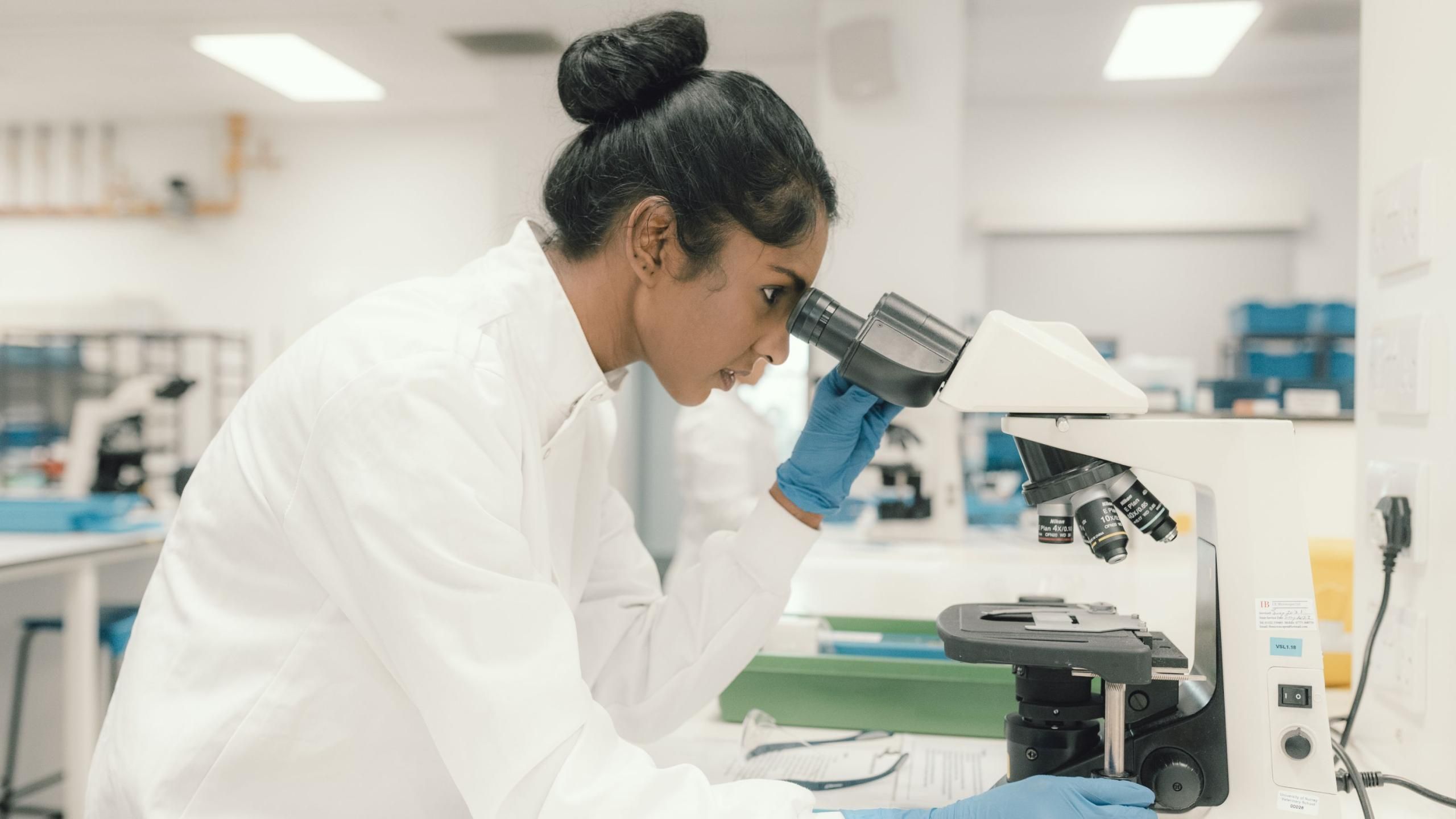 A woman iwearing a white lab coat looking through a microscope