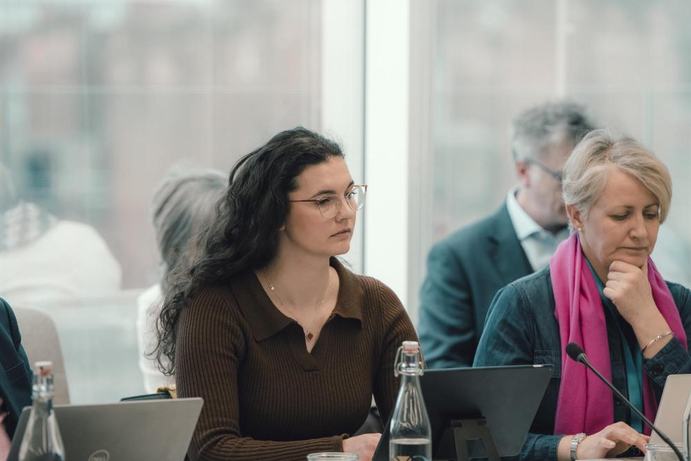 Photograph shows a professional meeting with attendees seated at a conference table, using laptops and microphones. 