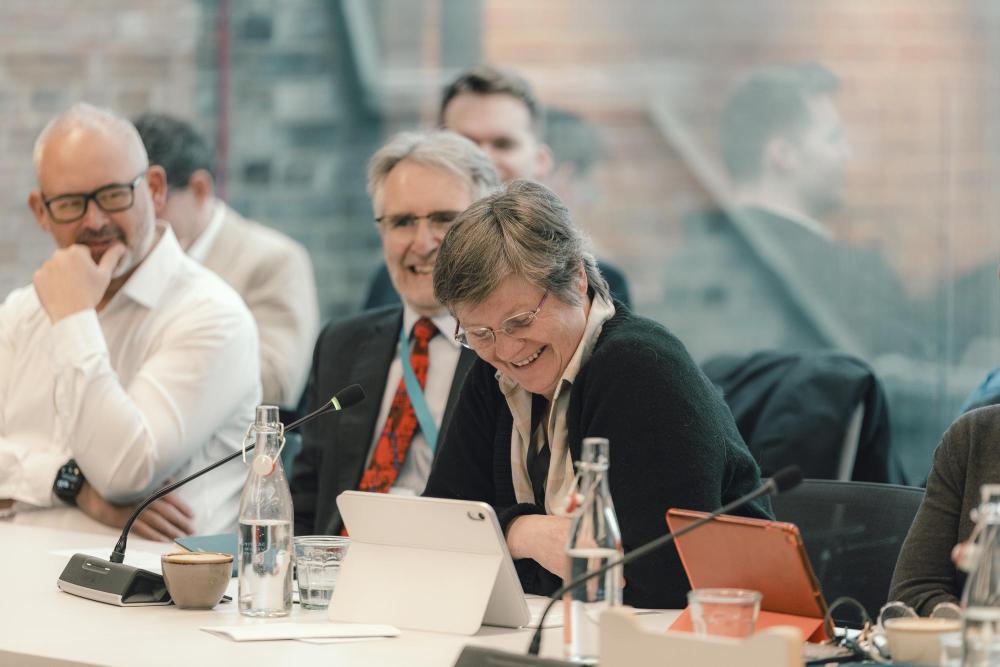 Photograph shows a group of professionals seated around a conference table during a meeting. 