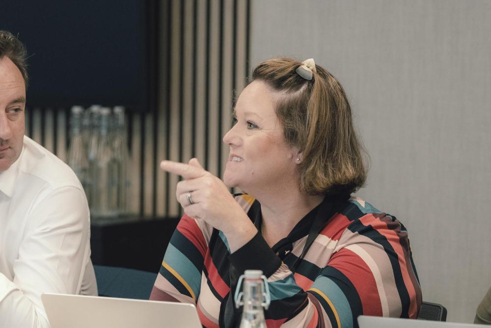 Photograph shows a woman speaking and pointing during a business meeting, with a partially visible man, laptop, and water bottle nearby.